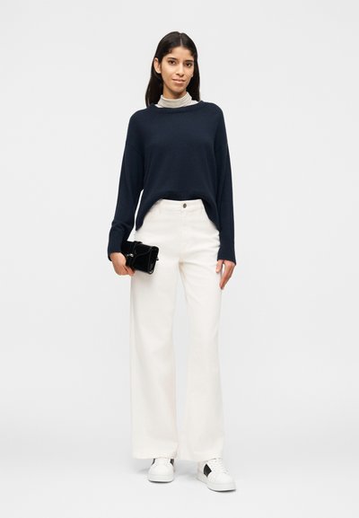 Γυναίκα που φοράει μπλε σκούρο πουλόβερ, λευκό παντελόνι με φαρδιά μπάτσες, λευκά αθλητικά παπούτσια, κρατώντας ένα μαύρο φάκελο σε άσπρο φόντο.