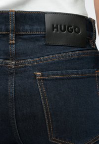 Vaqueros de mezclilla azul oscuro con una etiqueta de cuero negro en relieve con la palabra "HUGO" en la parte trasera, detallados con costuras naranjas y bolsillos clásicos.