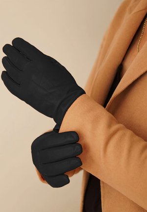 Czarne rękawiczki wykonane z miękkiego materiału, z przeszyciami i dopasowanym krojem, noszone na beżowym płaszczu.