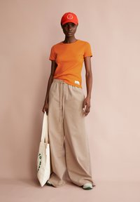 Oranje geribbelde T-shirt, beige broek met wijde pijpen, sneakers met groene accenten en een rode pet met wit logo. Katoenen draagtas met groene tekst.