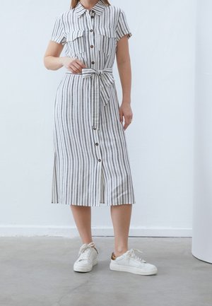 Femme portant une robe chemise midi blanche avec des rayures noires verticales, boutonnée sur le devant, taille ceinturée, et des baskets blanches avec des accents dorés.