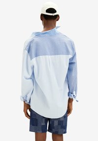 Hombre con una camisa de rayas de manga larga y suelta en tonos azules mezclados, pantalones cortos de estampado azul marino y una gorra de béisbol blanca, visto desde atrás.