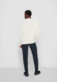 Weißer Strickpullover mit einem strukturierten Muster, kombiniert mit dunkelblauen, schmal geschnittenen Hosen und dunklen Schuhen. Besitzt einen Farbakzent am Halsauschnitt.