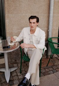 Modelul stă la o masă metalică rotundă, purtând o jachetă albă, pantaloni bej și pantofi negri, ținând o cană de cafea. Ochi de soare și un scaun verde sunt vizibile.