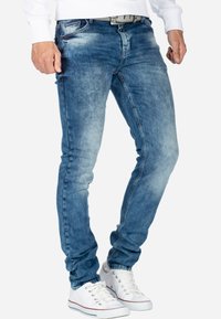 Jean en denim bleu délavé à coupe slim, avec une fermeture à bouton sur le devant et cinq poches, assortis à des baskets blanches.