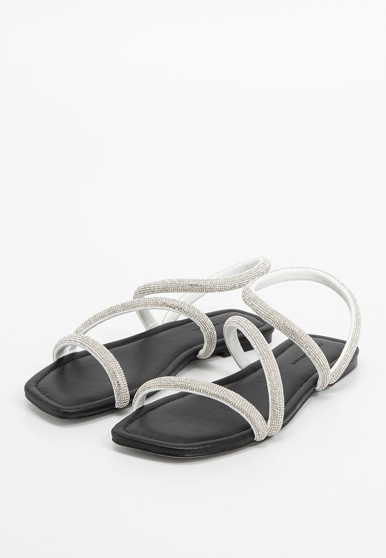 Platte sandalen met een zwarte zool. Zilveren banden met kralenaccenten vormen een gekruist ontwerp, wat zorgt voor een open teen-stijl.