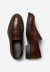 Brown leather loafers with a smooth texture, a flat rubber sole, and a front strap accent. Features cushioned insoles for comfort.