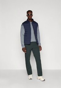 Marineblaue Steppweste über einem hellgrauen Hemd, kombiniert mit dunkelgrünen Cargohosen. Das Modell trägt weiße Turnschuhe und eine Sonnenbrille. Schlichter Hintergrund.