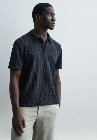 Un homme se tient de profil vers la droite, portant un polo noir texturé à manches courtes et un pantalon beige clair, devant un fond gris clair uni.