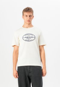 Jaunas vyras vilkintis off-white spalvos Jack & Jones marškinėliais su mėlynu ovaliu logotipu ir juodomis džinsomis, stovintis prieš paprastą baltą foną.
