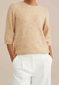 Beige stickad tröja med guldpaljetter, rund halsringning och trekvartsärmar, i kombination med vita plisserade byxor. Mjuk textur.