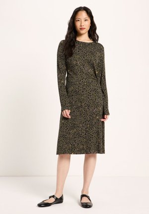 Woman wearing a knee-length olive green dress with a black and yellow leopard print and black flat Mary Jane shoes.