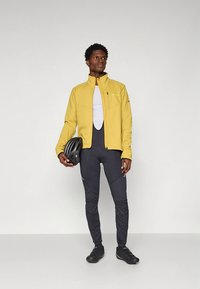 Yellow cycling jacket with a high collar, paired with black cycling pants. Model holds a black helmet, standing on a neutral background.