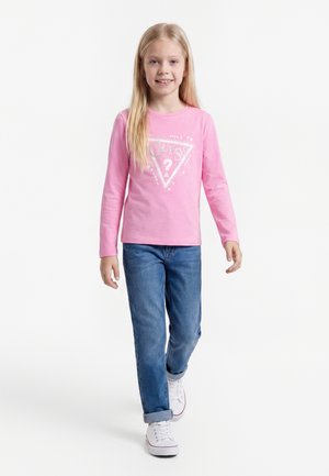 Jeune fille marchant, portant un t-shirt à manches longues rose avec un logo, un jean bleu avec les revers roulés et des baskets blanches sur un fond uni.