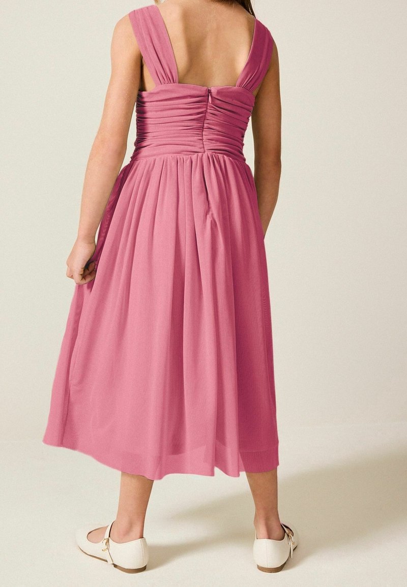 Fille portant une robe midi rose sans manches avec un dos froncé et des chaussures plates blanches, debout devant un fond clair uni.