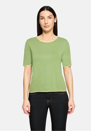 Kobieta o prostych ciemnych włosach, ubrana w jasnozieloną bluzkę z krótkim rękawem i ciemne dżinsy, stojąca na tle jednolitego jasnego tła.