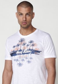 Baltos medvilnės marškiniai su grafika, kurioje pavaizduoti palmės mėlynos ir persikinės spalvos, su užrašu "Surf Rider" stilizuotu šriftu.