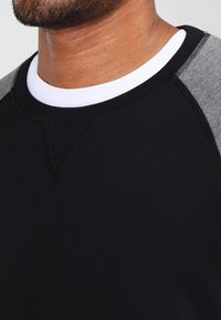 Svart sweatshirt med grå raglanärmar, rundad hals och en texturerad tygfinish med synliga sömnadsdetaljer.