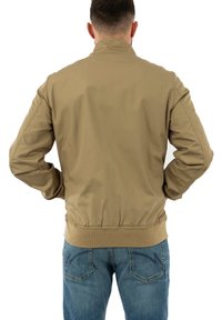 Veste bomber légère de couleur brun clair avec un col, une taille élastique et des poignets côtelés, associée à un jean bleu. Pas de logos ou de motifs visibles.