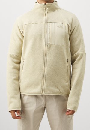 Homme portant une veste polaire beige zippée intégralement avec des poches latérales zippées et une poche poitrine sur un pantalon clair, devant un fond uni.