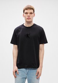 Melna kokvilnas t-krekla ar daļēji izšūtu CK logo uz krūtīm, apaļa kakla izgriezums un īsas piedurknes. Valkāts ar gaiši ziliem džinsiem.
