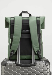 Grön tygryggsäck med rulltoppsdesign, vadderade axelremmar, mesh-bak och fack med dragkedja, vilande på en silverfärgad resväska med hexagonmönster.