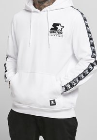 Felpa bianca con logo nero Starter sul petto, nastro con logo nero lungo le maniche, tasca frontale, indossata con le mani in tasca e pantaloni neri.