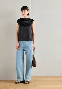 Top senza maniche in satin nero con scollatura scolpita, abbinato a jeans a gamba larga blu chiaro. Il modello tiene in mano una borsa in pelle marrone e indossa sandali neri.
