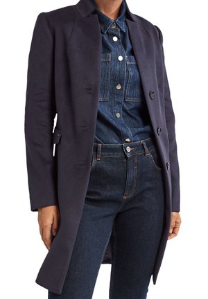 Osoba w ciemnogranatowym, dopasowanym płaszczu na guziki, na koszuli jeansowej i ciemnoniebieskich dżinsach, stojąca z rękami luźno opartymi po bokach.