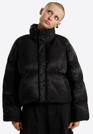 Person mit rasiertem Kopf, trägt eine schwarze übergroße Steppjacke und schwarze Hose, neigt den Kopf leicht nach oben und zur Seite.