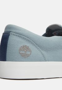 Zapatillas de lona azules con un talón texturizado en azul oscuro, suela blanca y un logo de árbol bordado en gris en el lateral.