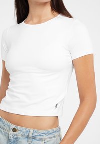 Biała krótka bluzka z krótkim rękawem wykonana z gładkiego, elastycznego materiału. Posiada okrągły dekolt oraz małe czarne logo po dolnej stronie.