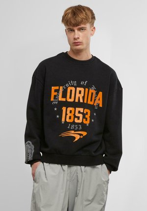 Sort sweatshirt med "Florida 1853" i fed orange og grå tekst, med stjernesymboler og en detaljeret grafik på ærmet.