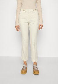 Pantalon blanc sur mesure à coupe slim avec des accessoires dorés élégants à la taille. Associé à des chaussures compensées marron épaisses et un haut beige.