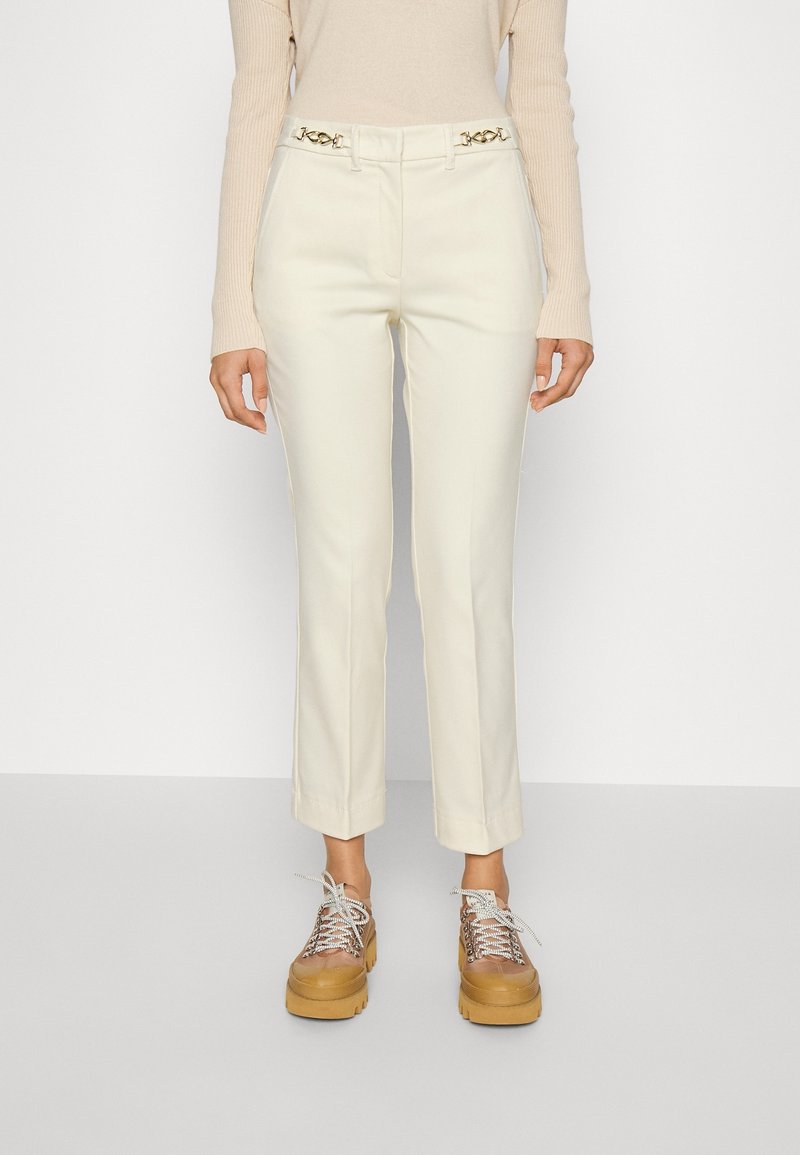 Pantalon blanc sur mesure à coupe slim avec des accessoires dorés élégants à la taille. Associé à des chaussures compensées marron épaisses et un haut beige.