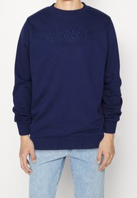 Uomo che indossa una felpa blu navy con ricamato sul petto "Lyle & Scott Established 1874" e jeans azzurri.
