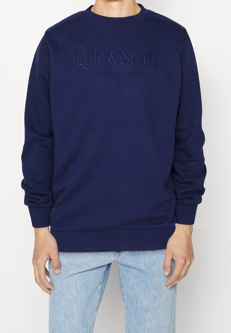Uomo che indossa una felpa blu navy con ricamato sul petto "Lyle & Scott Established 1874" e jeans azzurri.