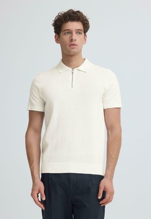 Nooruk lokkis juustega, kes kannab valget lühikese varrukaga polosärki ees lukuga ja tumedaid pükse, seisab ühevärvilise tausta ees.