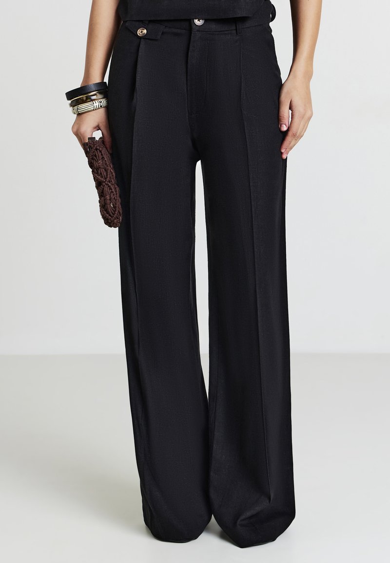 Pantalons larges noirs en tissu texturé avec deux poches avant, accentués par deux boutons, associés à une pochette à motif marron.