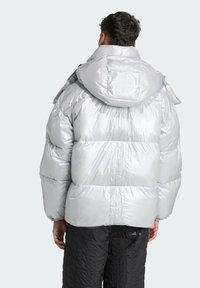 Strieborná nafukovacia bunda s lesklým povrchom, vybavená kapucňou, prešívanými panelmi a elastickými šnúrkami na stiahnutie. Nosená na čiernych textúrovaných nohaviciach.