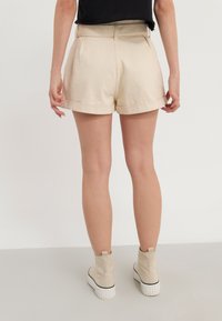 Beige kraťasy s ohrnutými manžetami, se bočními kapsami a hladkým, strukturovaným materiálem. V kombinaci s béžovými kotníkovými botami s bílou podrážkou.