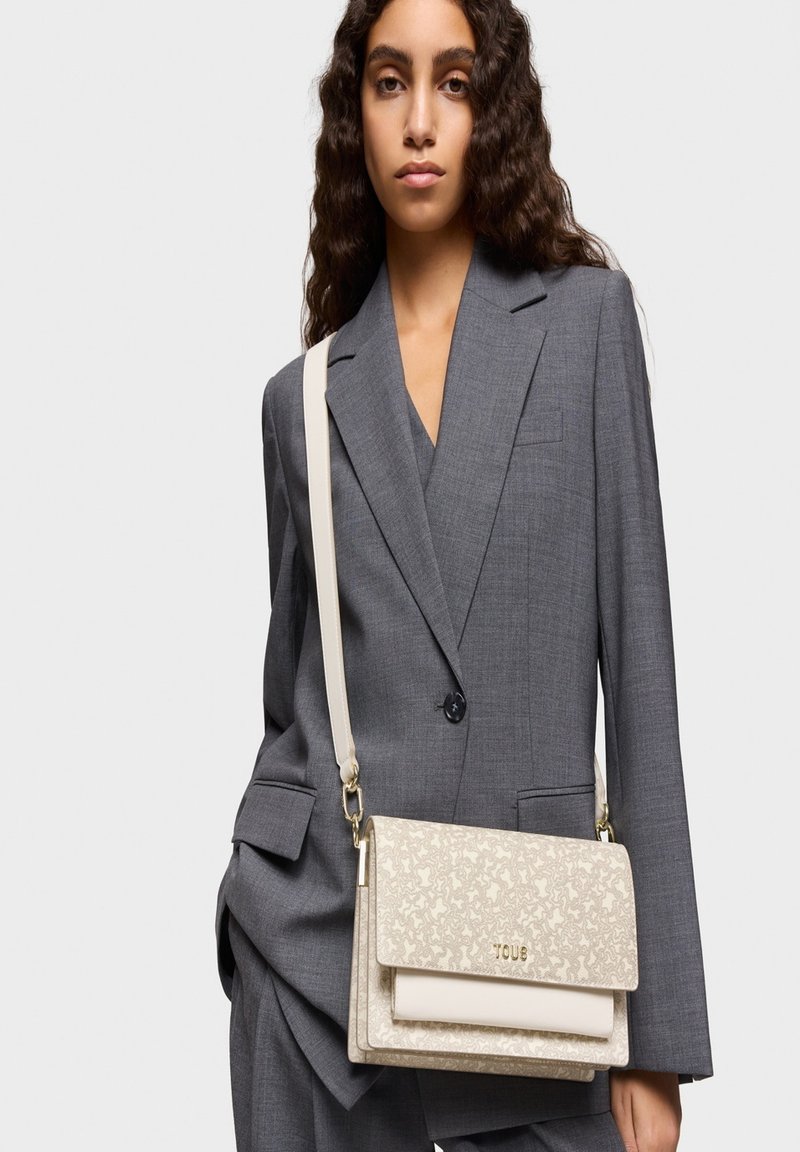 Bolso de mano beige con patrón y logo en tono dorado, que presenta un diseño estructurado y una correa de hombro ajustable, combinado con un traje gris.