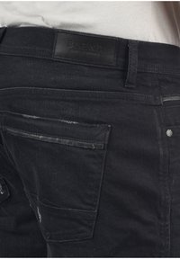 Svarta denimjeans med en strukturerad yta, som har en subtil slitdetalj, bakficka med en elegant svart kant och en läderloggopatch.