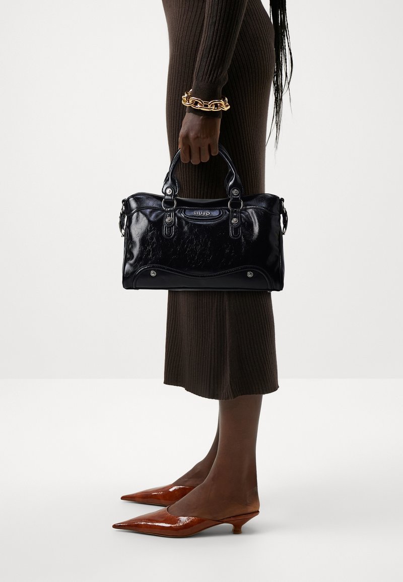 Personne tenant un sac à main noir brillant, portant une robe côtelée marron, un bracelet chaîne en or et des chaussures marron pointues à talon bas sur un fond uni.