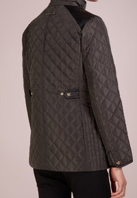 Vadderad olivgrön jacka med hög krage, svarta läderaxlar och guldfärgade detaljer på baksidan och ärmarna.