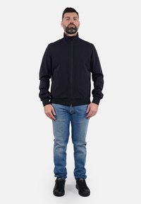 Giacca nera con zip e colletto alto, due tasche anteriori e polsini elasticizzati, abbinata a jeans blu e sneaker nere.