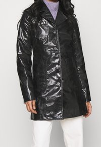 Manteau noir brillant à coupe ajustée, fermeture à boutons, poches à rabat et col cranté. Fabriqué en matériau brillant, design mi-longueur.