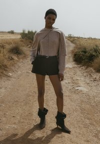 Stickad beige tröja med knappdetaljer, kombinerad med högmidjade svarta shorts och svarta slappa ankelstövlar. Dammig utomhusmiljö.