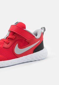 Scarpa sportiva rossa con tomaia in mesh texturizzato, accenti grigi, tallone nero e suola in gomma bianca. Presenta una cintura regolabile e lacci.