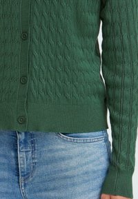Cardigan vert en tricot avec un motif de câble texturé, fermeture à boutons et bords côtes, associé à un jean en denim bleu clair.
